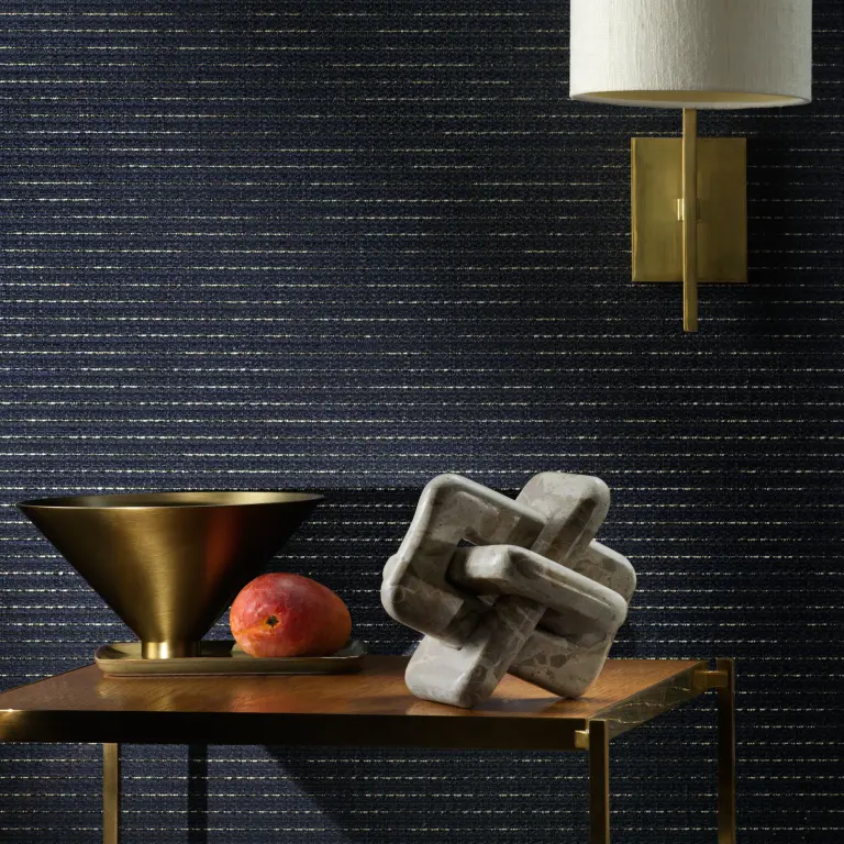 escom Eldor textile wallcovering with a refined woven structure and subtle metallic thread, creating a softly radiant surface, shown on interior walls in a styled setting with decorative objects and wall lighting