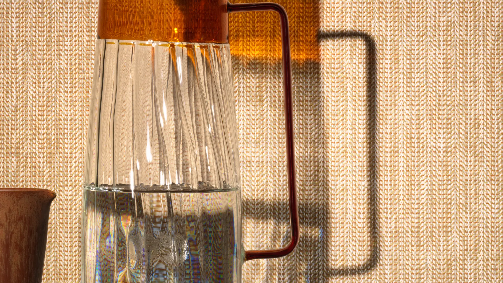 Vescom Sisu textile wallcovering with a structured herringbone weave, creating a measured and rhythmic surface, shown on interior walls in a styled setting with a glass carafe