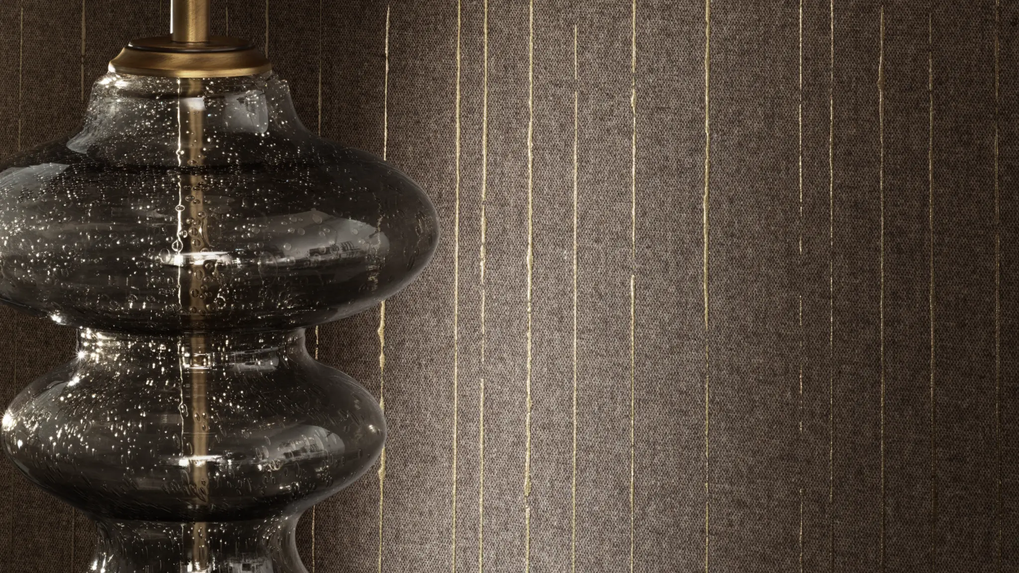 Vescom Sofia textile wallcovering with a warm woollen weave and subtle metallic threads creating a refined shimmer, shown on interior walls behind a decorative glass table lamp